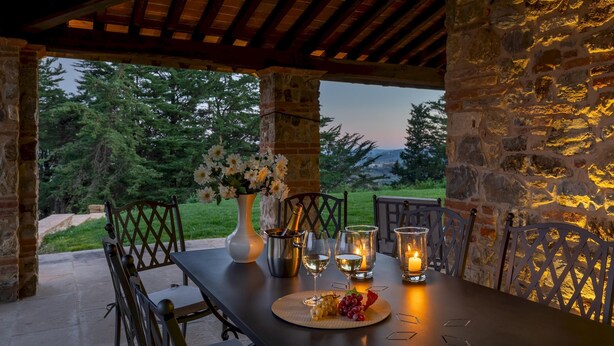 An outdoor dining area with a stone wall, wooden chairs, a table set with wine glasses, a bottle of champagne, and a vase of flowers. - Villa Dada 10 Emma Villas (Rosignano Marittimo)