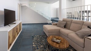 A modern living room with a beige sofa, a wooden coffee table, and a flat-screen TV on a white console. A staircase with glass railings leads to an upper level. - Villa Ristola 6 Emma Villas (Castrignano del Capo)