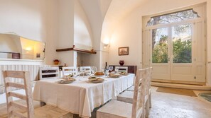 Un espace repas avec une longue table dressée pour un repas, des chaises blanches et une grande fenêtre donnant sur un paysage verdoyant.