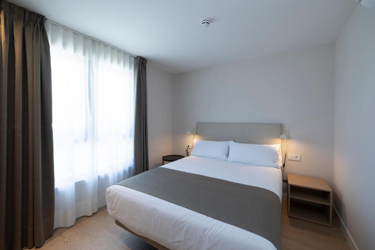 A modern hotel room with a large bed, a bedside table, a nightstand, and a window with curtains.