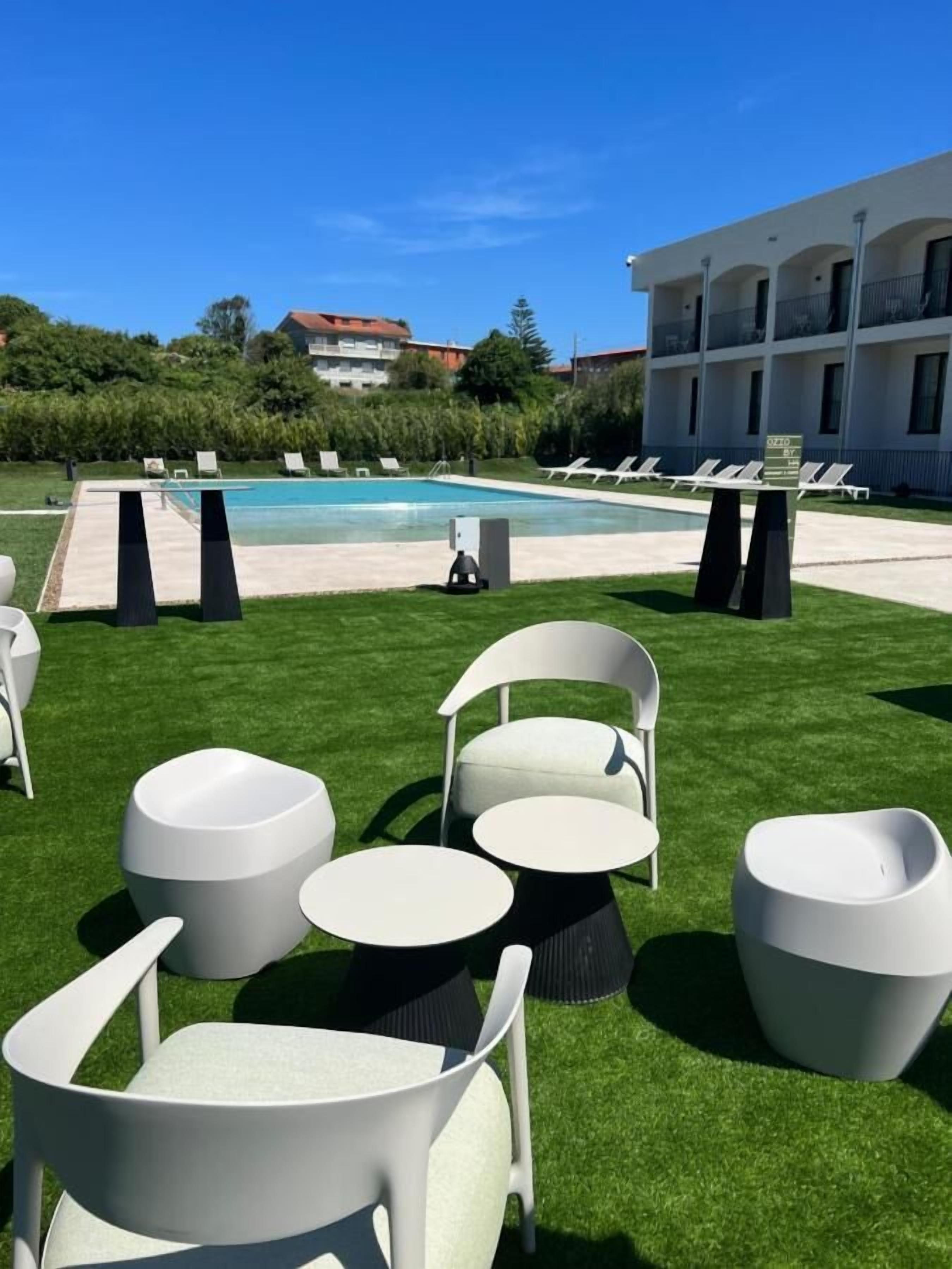 Un área junto a la piscina con sillas y mesas de jardín blancas, cielo azul despejado y un edificio al fondo.