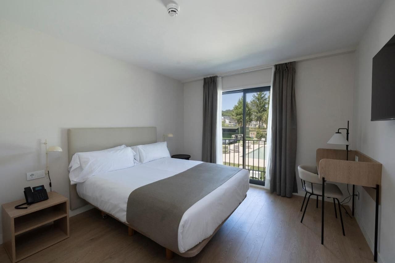 Una habitación de hotel con cama, escritorio, silla, teléfono y ventana con cortinas.