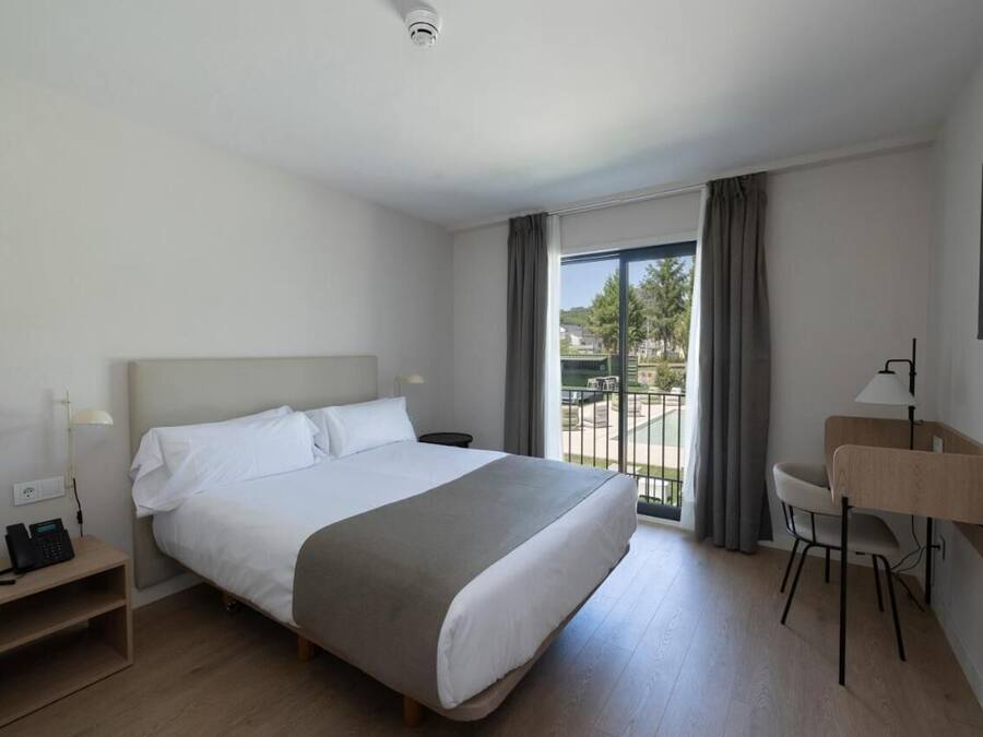 Una habitación de hotel con cama, escritorio, silla, teléfono y ventana con cortinas.