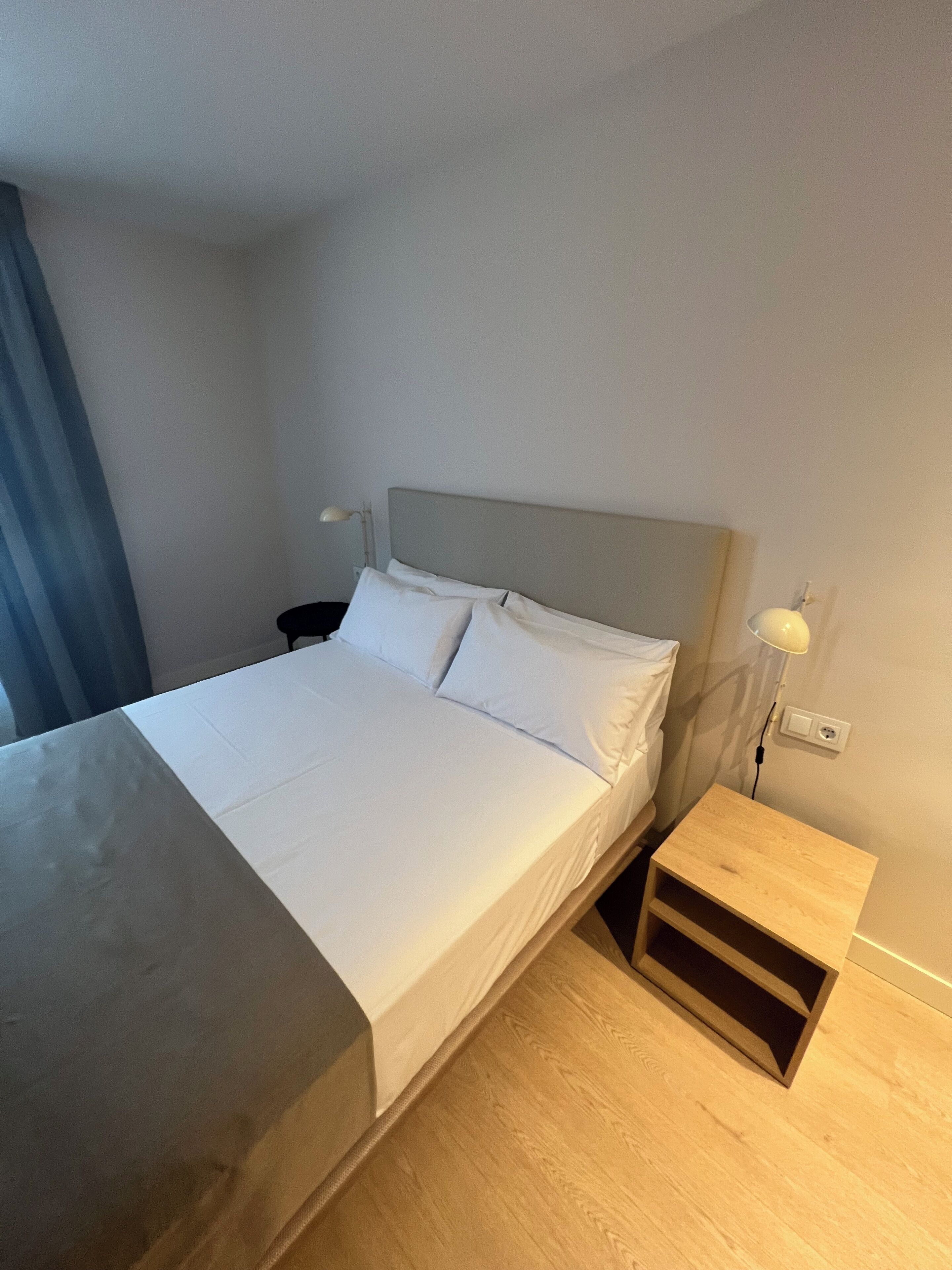 Una cama bien hecha con ropa de cama blanca, una mesita de noche con lámpara y un enchufe en la pared.