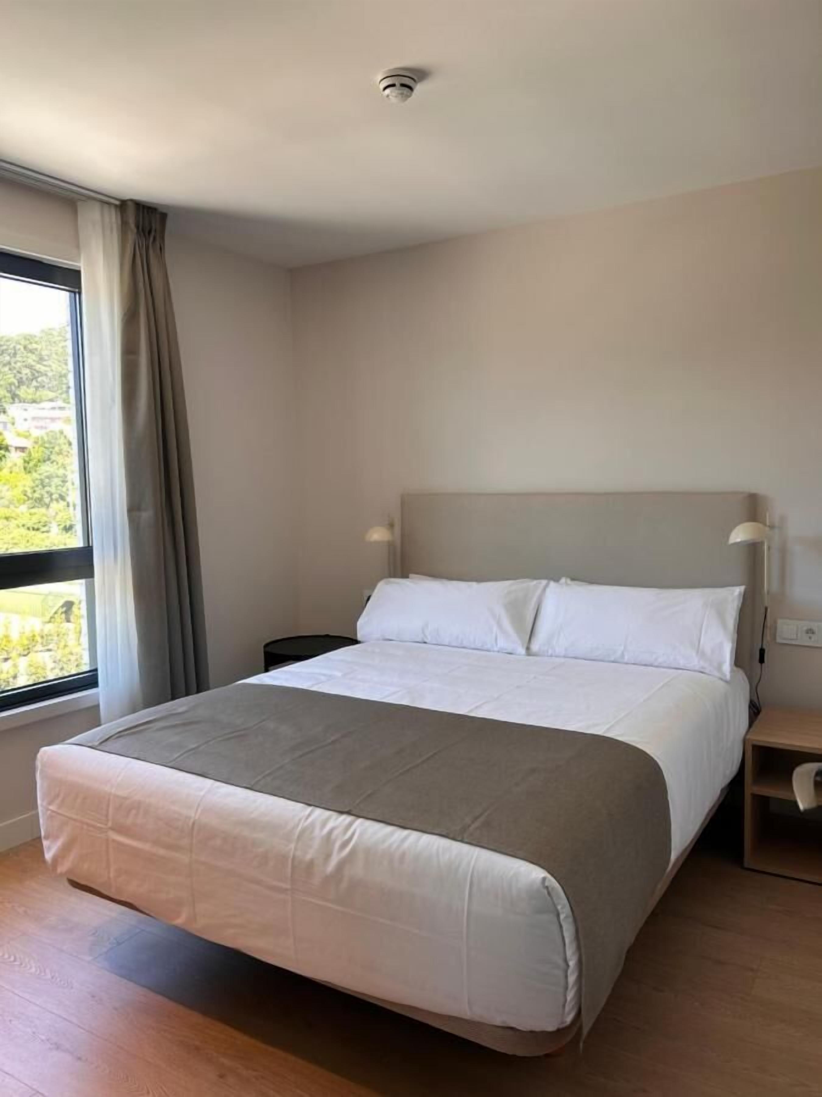 A modern hotel room with a large bed, a nightstand, a lamp, and a window with curtains.