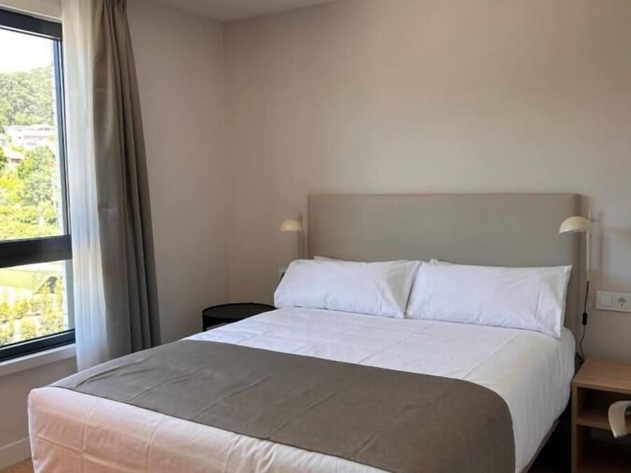 A modern hotel room with a large bed, a nightstand, a lamp, and a window with curtains.