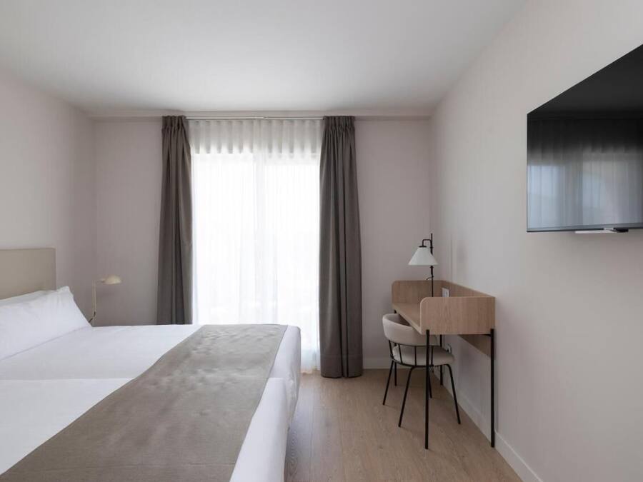 A hotel room with a bed, a desk, a chair, a TV, and a window with curtains.