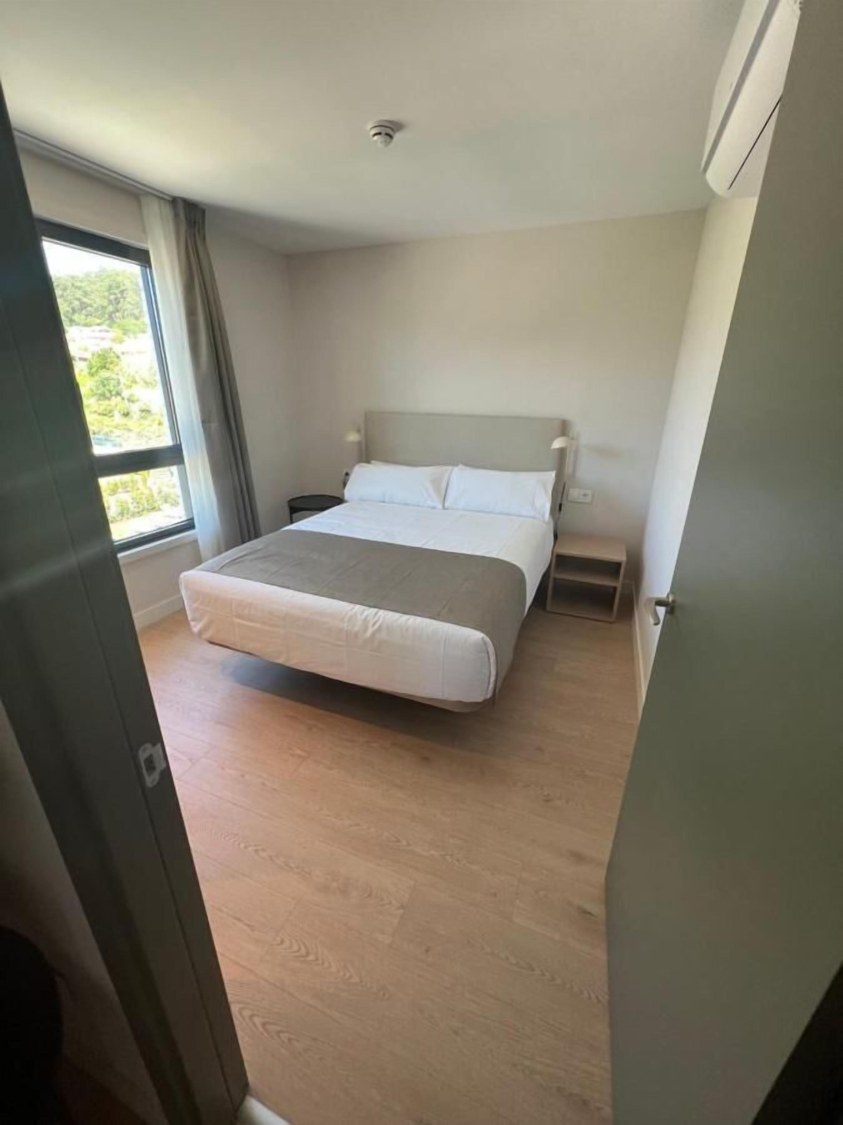 Un dormitorio con cama, mesitas de noche, una ventana y un aire acondicionado instalado en el techo.
