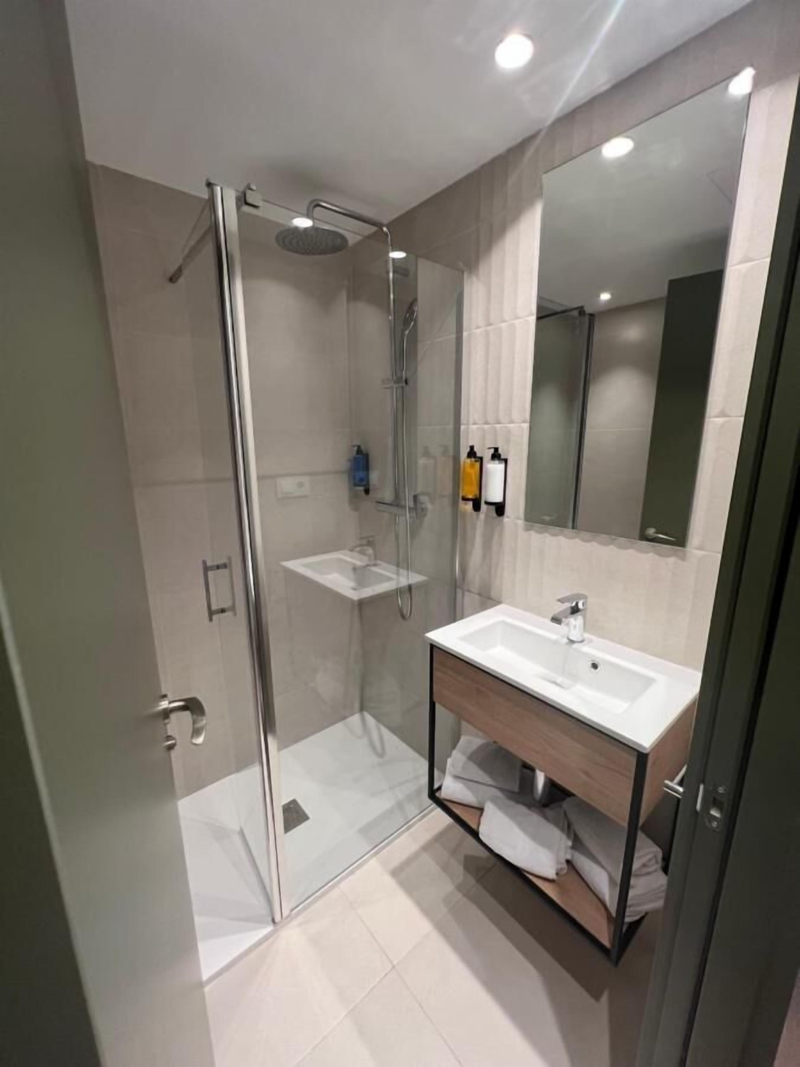 Un baño moderno con ducha envuelta en vidrio, un lavamanos blanco y un mueble de tocador de madera.