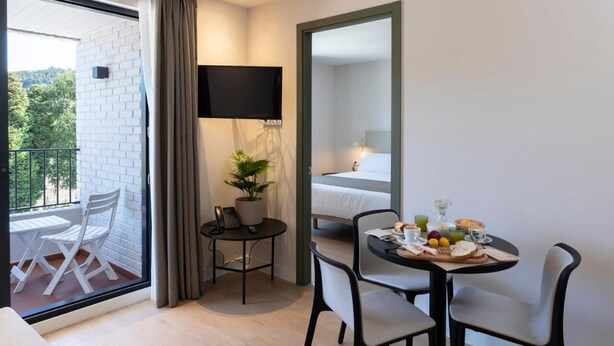 A hotel room with a balcony, a dining area with a table set for breakfast, a flat-screen TV, and a bed with a white headboard. - Hotel H4 Cangas (Cangas)