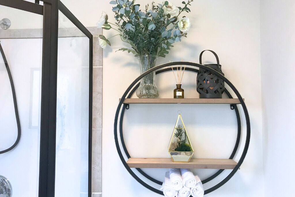 Un baño con cabina de ducha de vidrio, un estante en la pared con objetos decorativos y un perchero con toallas.