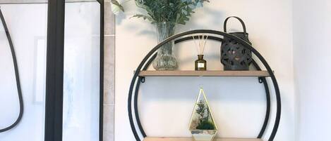 Un baño con cabina de ducha de vidrio, un estante en la pared con objetos decorativos y un perchero con toallas.