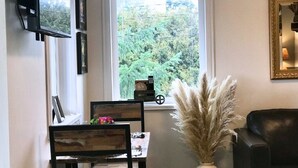 A room with a dining table, chairs, a vase with pampas grass, a black leather sofa, and a window with a view of greenery.