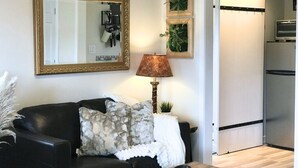 A living room with a black leather sofa, a wooden coffee table, a gold-framed mirror, a wall-mounted lamp, and a kitchen area with a refrigerator and a microwave.