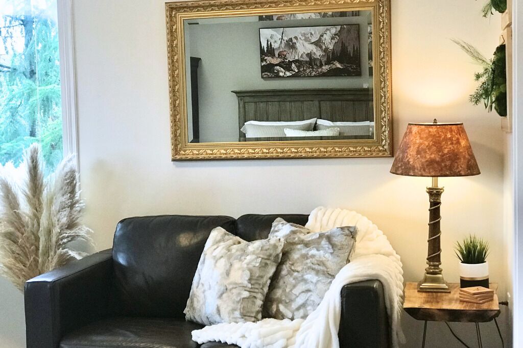 Un sofá de cuero negro con una manta blanca y almohadones decorativos, un espejo con marco dorado arriba y una lámpara de mesa a la derecha.