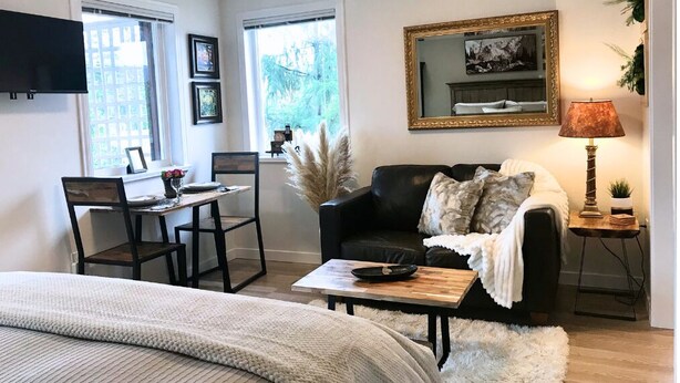 A modern living room with a black leather sofa, a wooden coffee table, a white fluffy rug, a dining table with chairs, and a TV mounted on the wall.