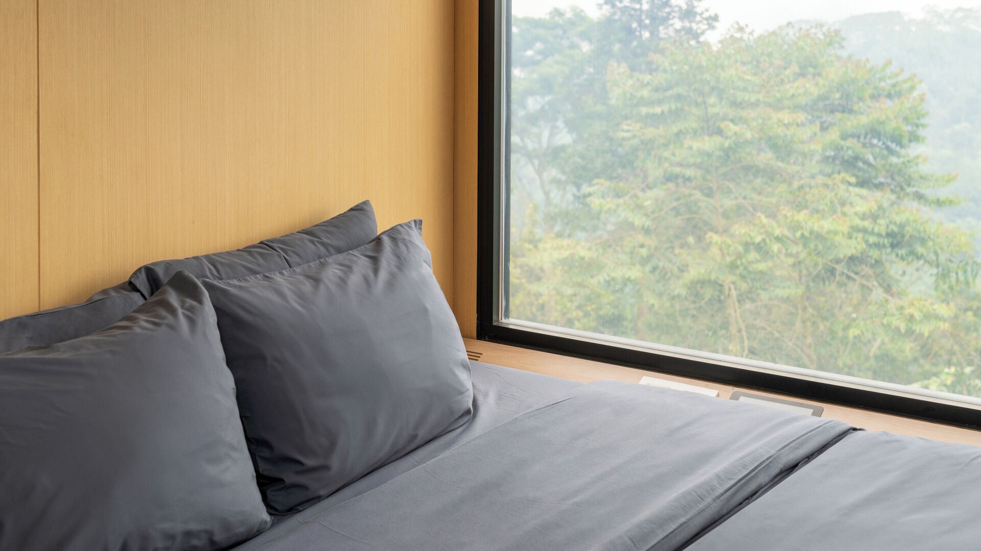 Ein Schlafzimmer mit großem Fenster, Holz-Kopfteil und grauer Bettwäsche.
