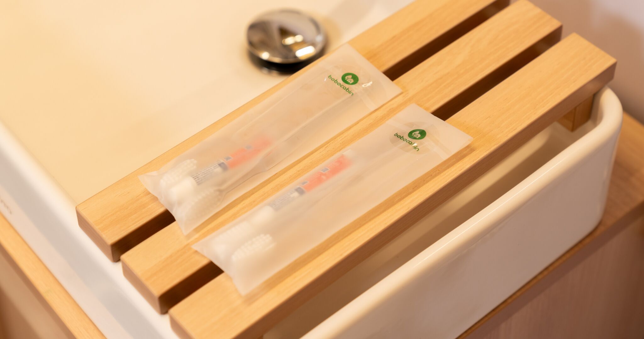 Una bandeja de madera con dos bolsas de plástico transparentes que contienen artículos de aseo sobre un lavamanos blanco.
