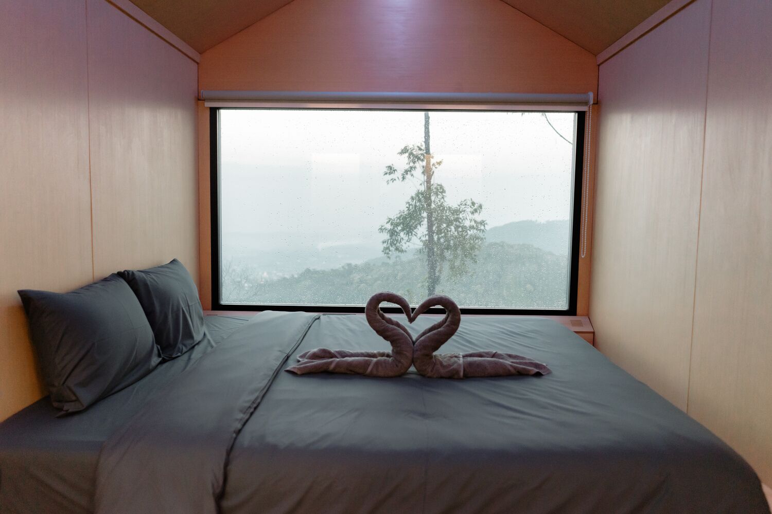 Une chambre avec une grande fenêtre, un lit avec une literie grise et une décoration en forme de cœur réalisée avec des serviettes.