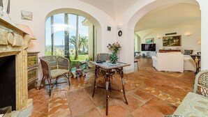 Un salón amplio con chimenea, sillas de mimbre, una mesa de centro y vistas a palmeras.