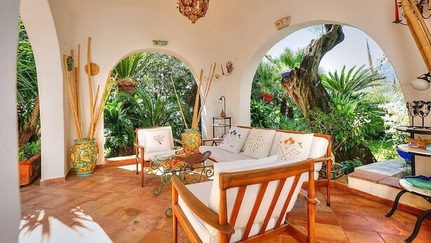 Un patio iluminado por el sol con un sofá de madera, una mesa y vista a un gran árbol a través de un arco.