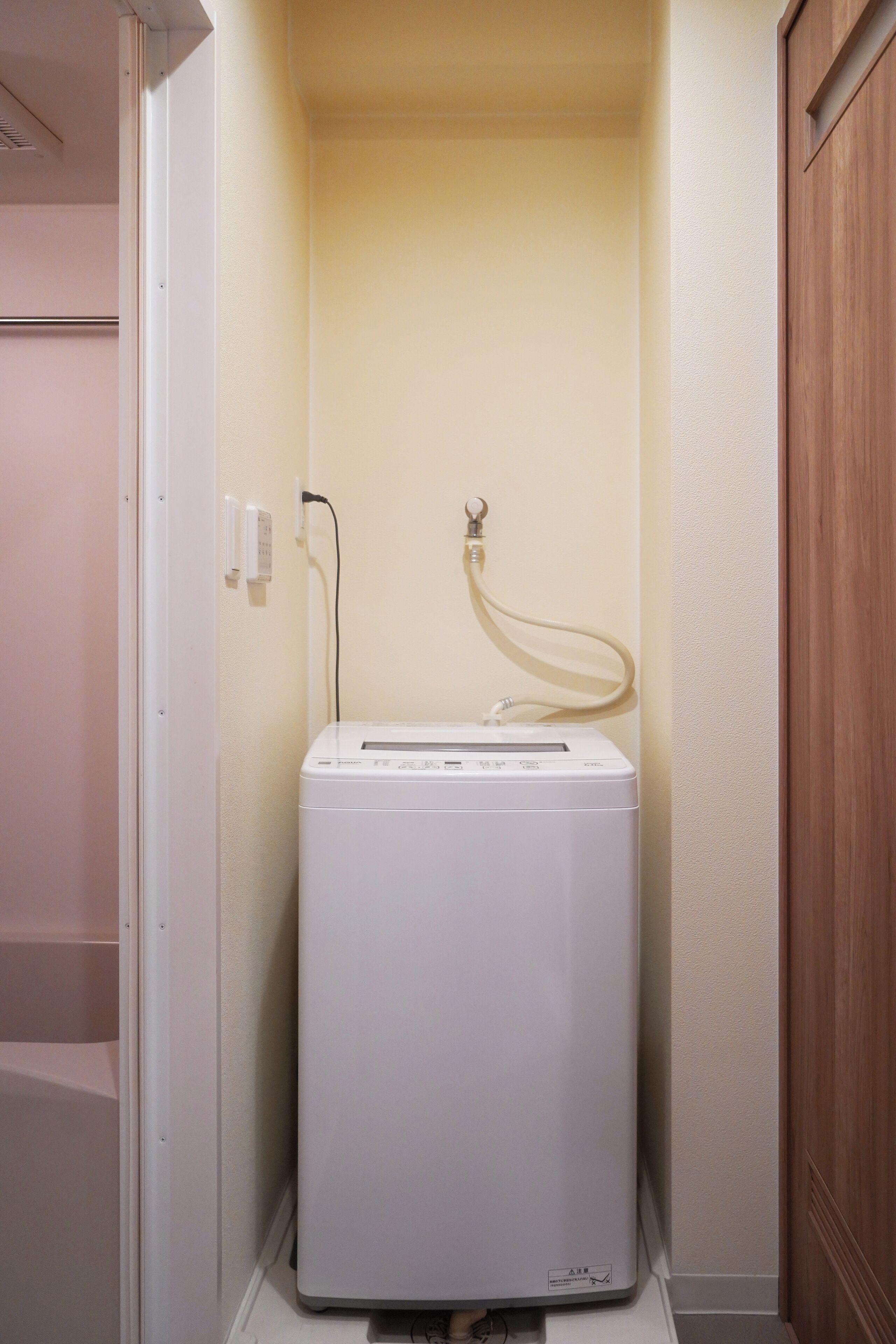 A compact, white, wall-mounted washer and dryer unit in a small, enclosed laundry room with a wooden door and a light-colored wall.