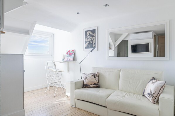 A room with a white sofa, a desk with a chair, a window, and a mounted TV.