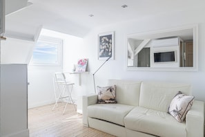A room with a white sofa, a desk with a chair, a window, and a mounted TV.