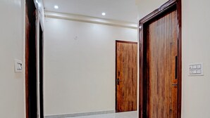 A hallway with white walls and a tiled floor, leading to two wooden doors. - Kashvi Residency (Greater Noida)
