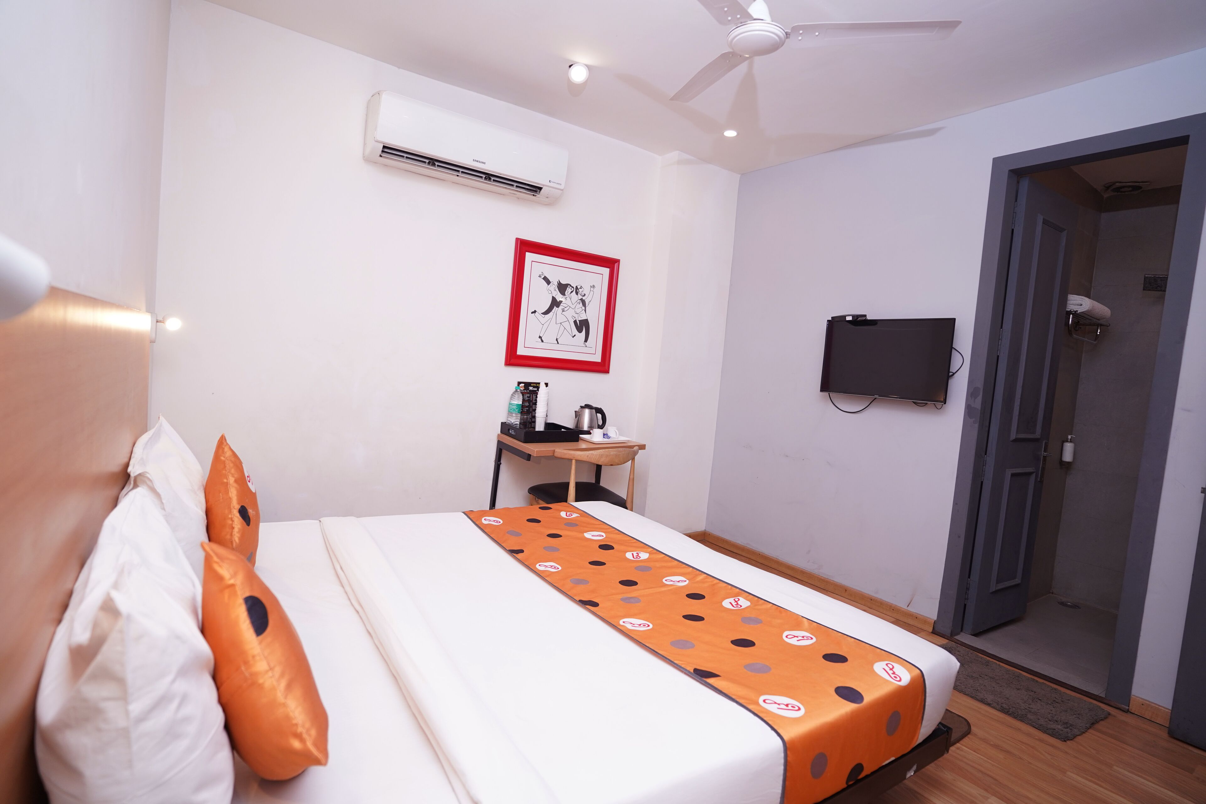 A hotel room with a bed, an air conditioner, a framed picture, a desk with a television, and a shower area.