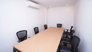 Una sala de conferències amb una taula de fusta llarga, sis cadires de despatx negres, parets blanques i una unitat de climatització al sostre.