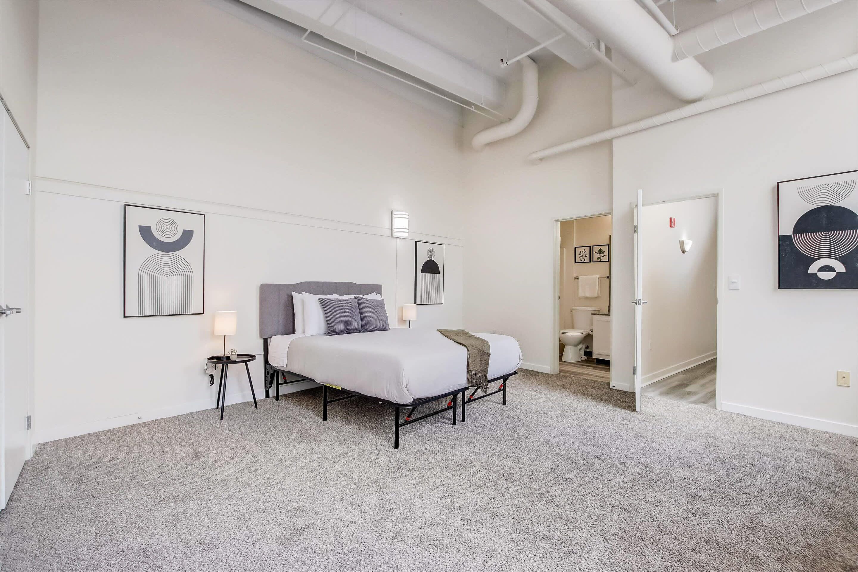 Un dormitorio moderno con una cama grande, una mesita de noche y un velador. Hay dos luces fijadas a la pared y una puerta que da a otra habitación.