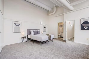 Une chambre moderne avec un grand lit, une table de chevet et une commode. On y trouve deux appliques murales et une porte qui donne sur une autre pièce.