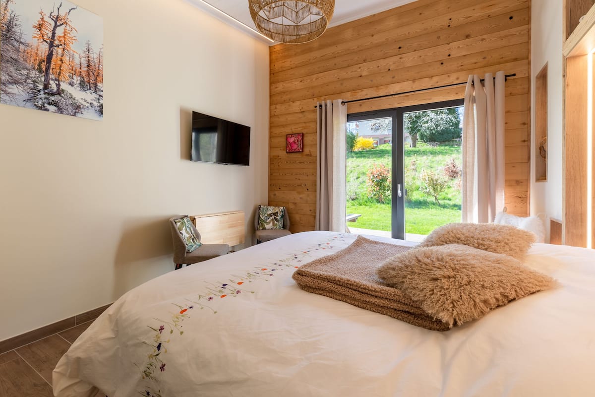 A bedroom with a large bed, wooden ceiling, a TV mounted on the wall, and a sliding glass door leading to a garden.