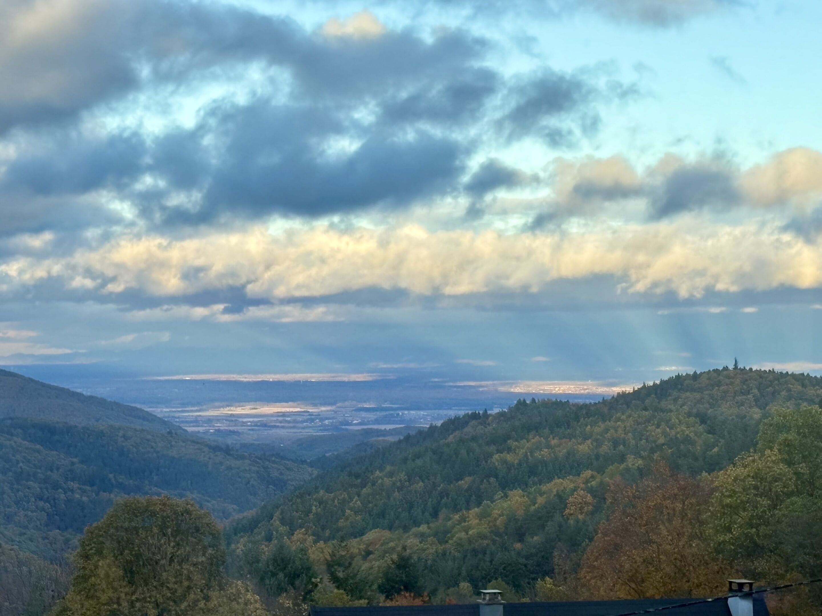 Un paysage montagneux avec une forêt dense, une vallée et, au loin, une vue sur une ville, le tout sous un ciel nuageux.