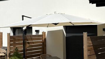 Un patio al aire libre con una mesa y sillas bajo un paraguas blanco, cerca de una valla de madera y una maceta con un árbol pequeño.