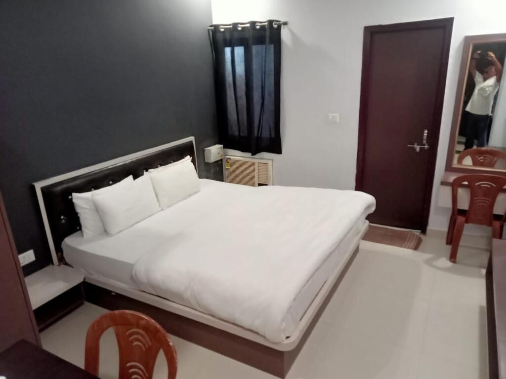 A neatly made bed in a hotel room with a dark wall, a window with curtains, a wooden door, and a small table with a chair.