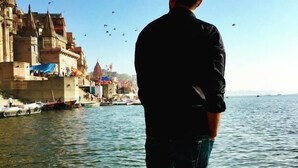 A person standing on a wooden boat facing a river with buildings on the opposite bank. - The White Canyon (Varanasi)