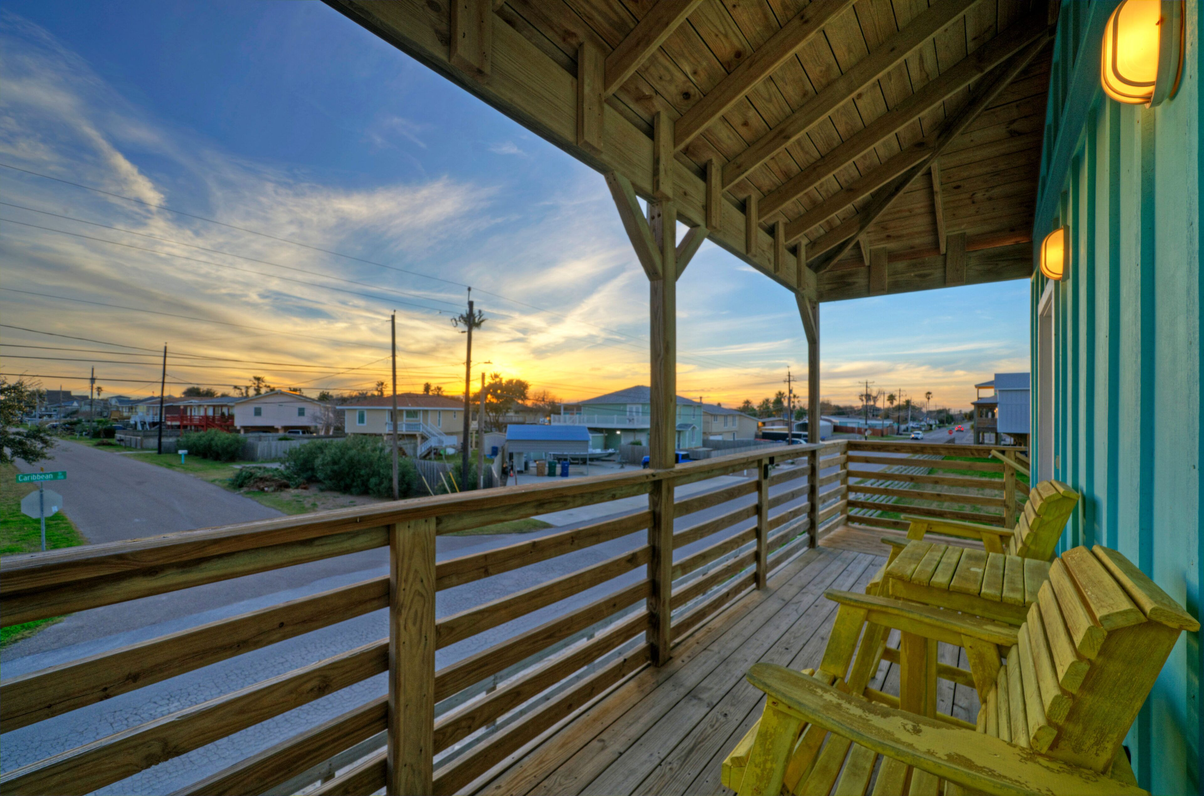 Una terraza de madera con barandilla que da vista a una calle y edificios, con un atardecer de fondo.