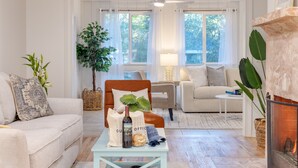 A cozy living room with a sofa, armchair, and coffee table. There is a fireplace, a plant, and a window with curtains. - Conch Cottage by AvantStay | Walk to Beach, Deck, Fireplace, Outdoor Dining (Isle Of Palms)