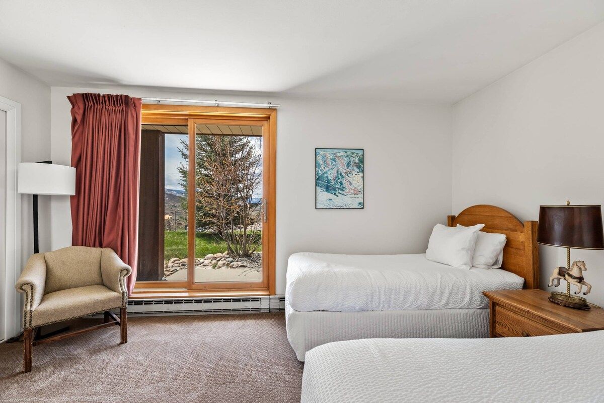 A hotel room with a bed, a chair, a nightstand, a lamp, a window with curtains, and a painting on the wall.