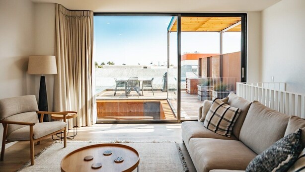 A modern living room with a sofa, armchair, and coffee table, a large sliding glass door leading to an outdoor patio with a wooden deck and seating area. - 95 Hardy Street Apartment 1 (Nelson)