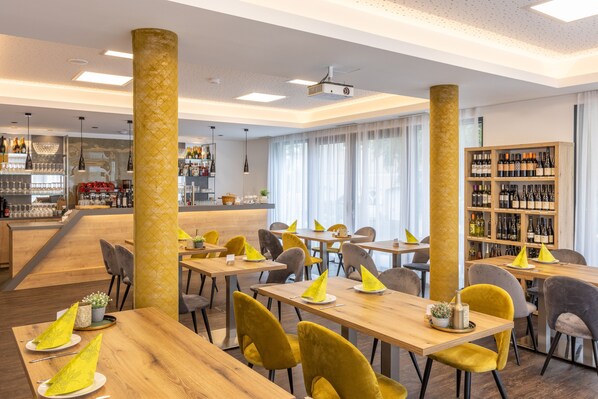 Un restaurante moderno con mesas de madera y sillas amarillas, zona de bar y una vitrina de vinos.