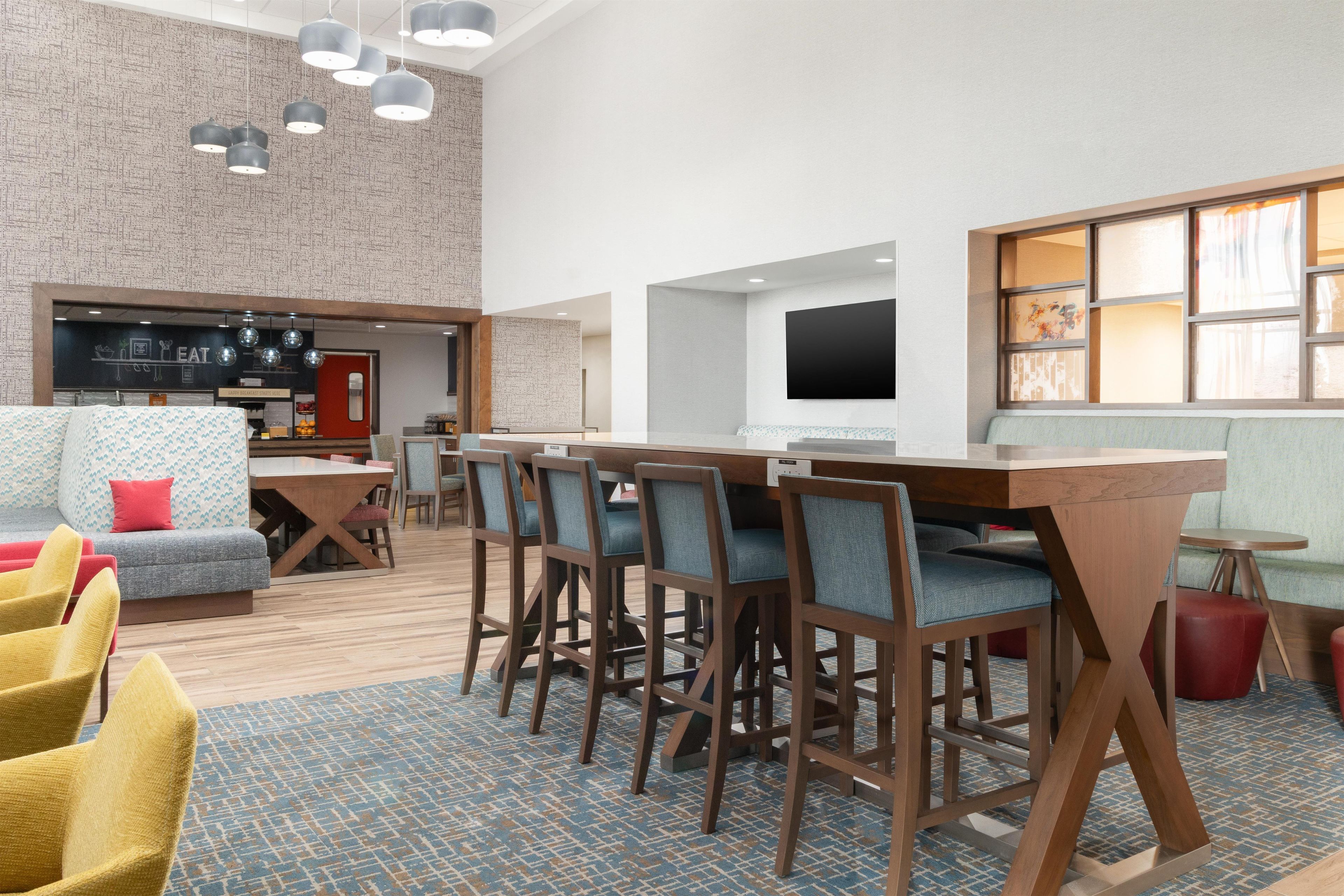 A modern hotel lobby with a bar area, seating arrangements, and a flat-screen TV mounted on the wall.