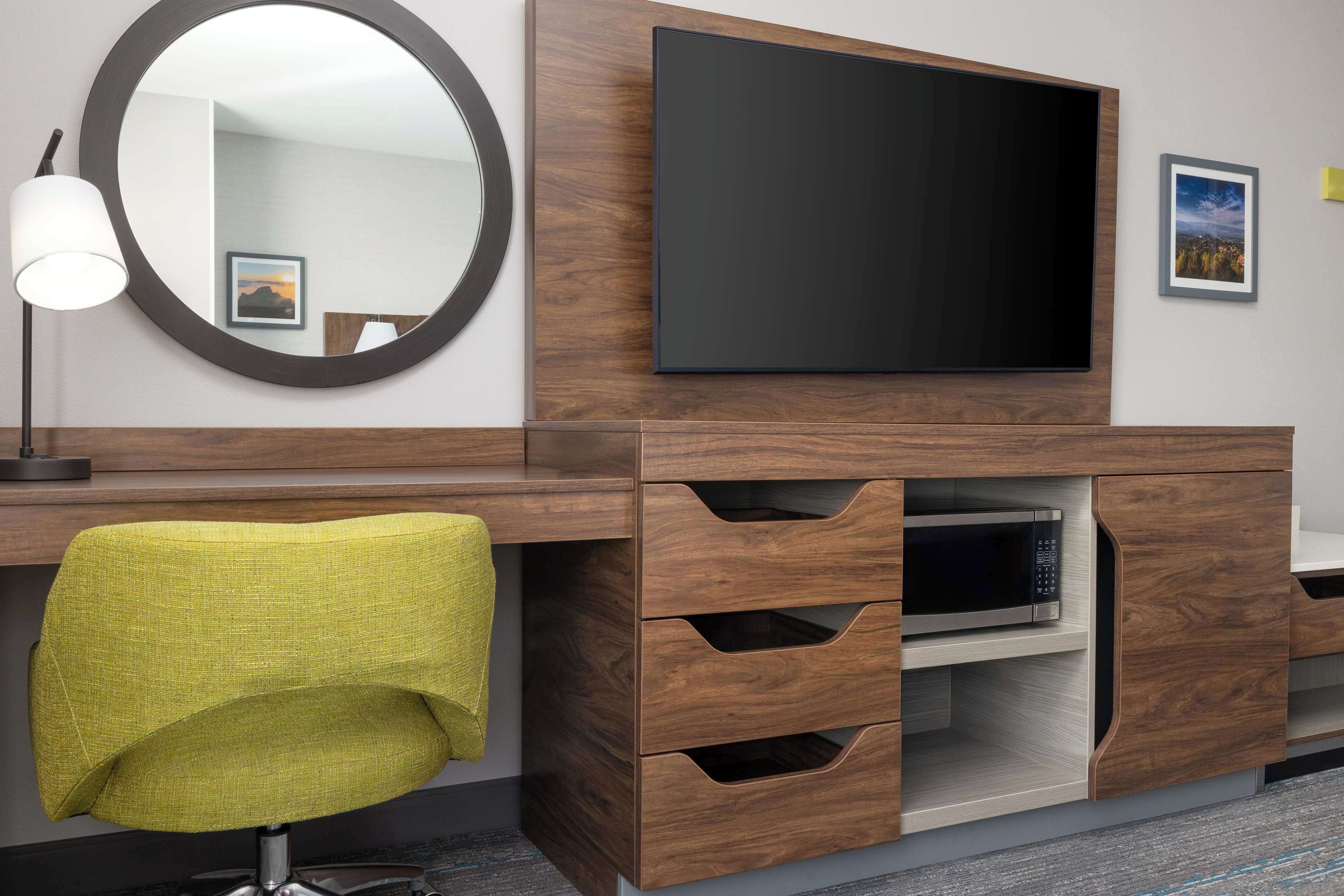 A hotel room with a wooden desk, a television, a mirror, and a green chair.