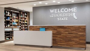A reception area with a "Welcome to the Golden State" sign, a wooden reception desk, and shelves stocked with snacks and toiletries. - Hampton Inn & Suites Porter Ranch Los Angeles (Porter Ranch)