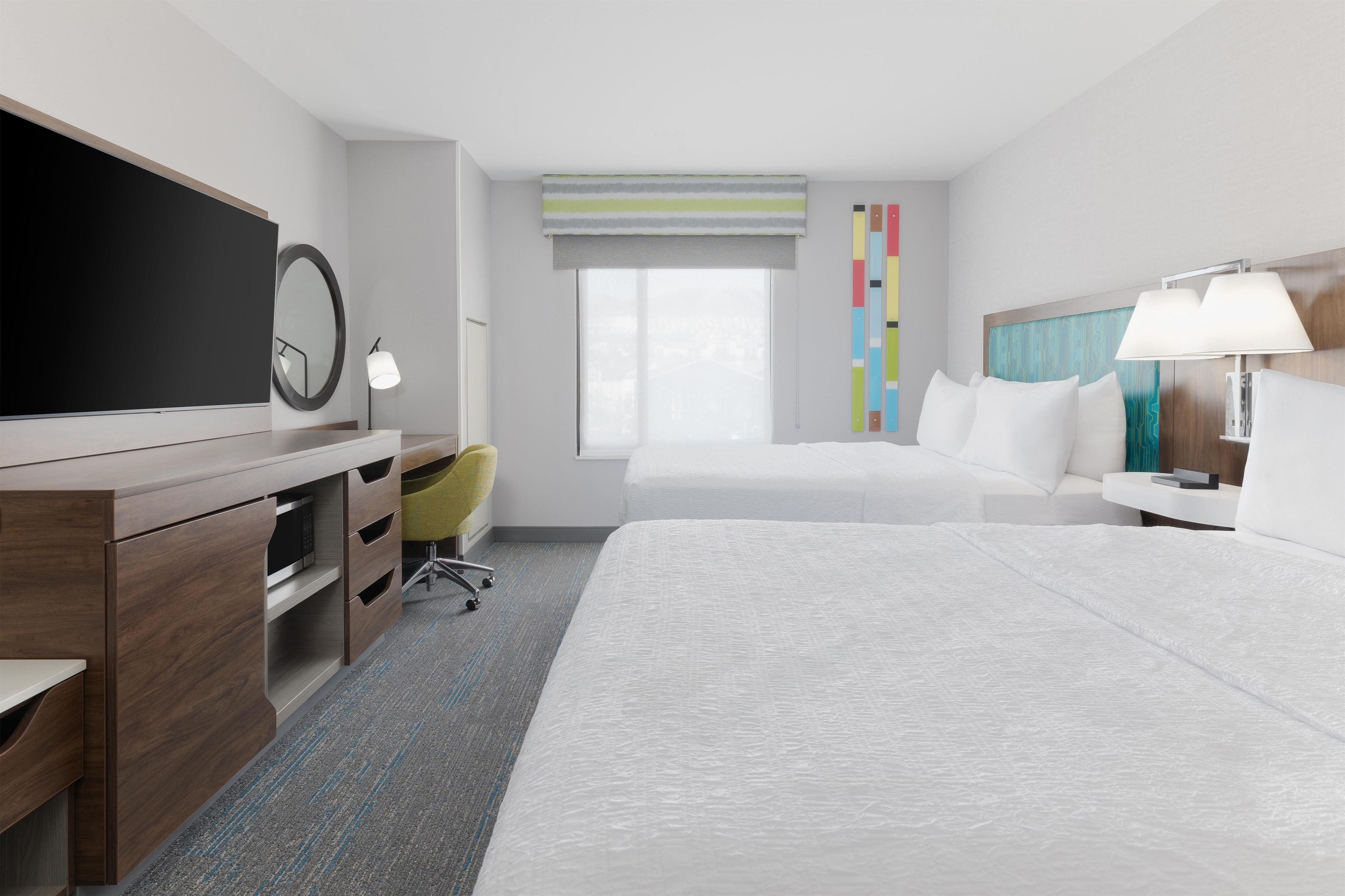 A hotel room with a large bed, a desk with a flat-screen TV, a chair, a bedside table with a lamp, and a window with a view.