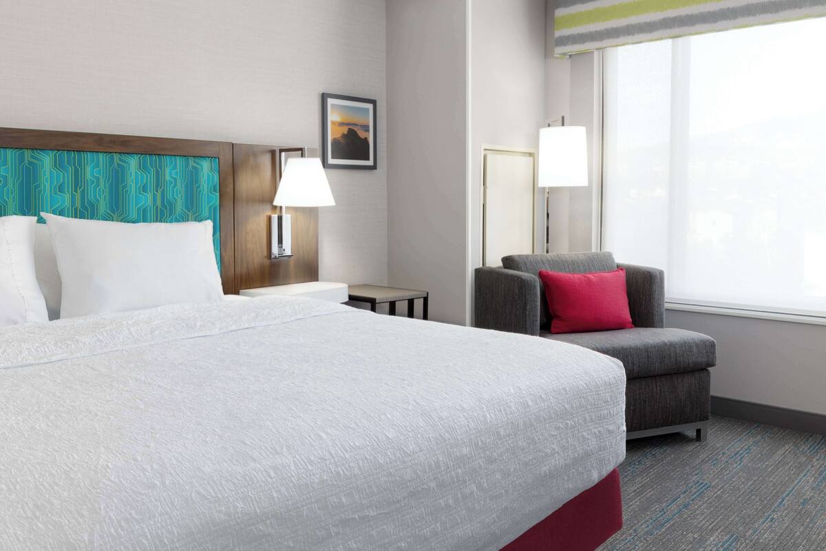 A hotel room with a large bed, a grey armchair with a red pillow, a nightstand with a lamp, and a picture on the wall.