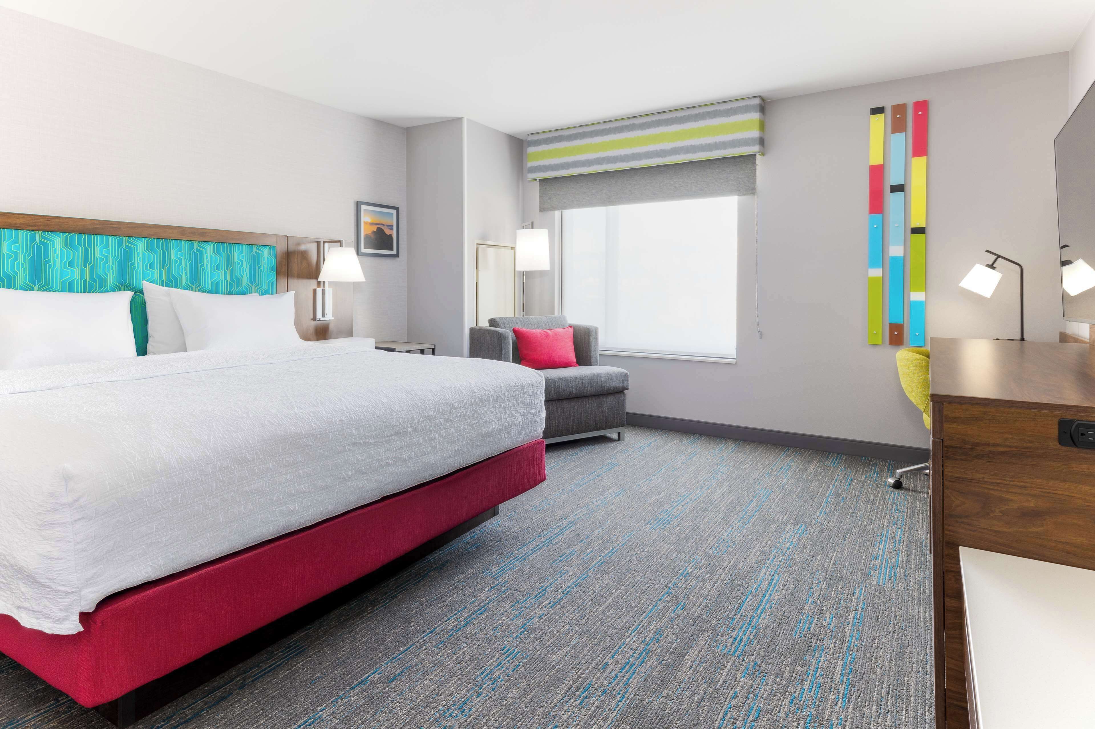 A modern hotel room with a large bed, a desk, a chair, and a window with a view.