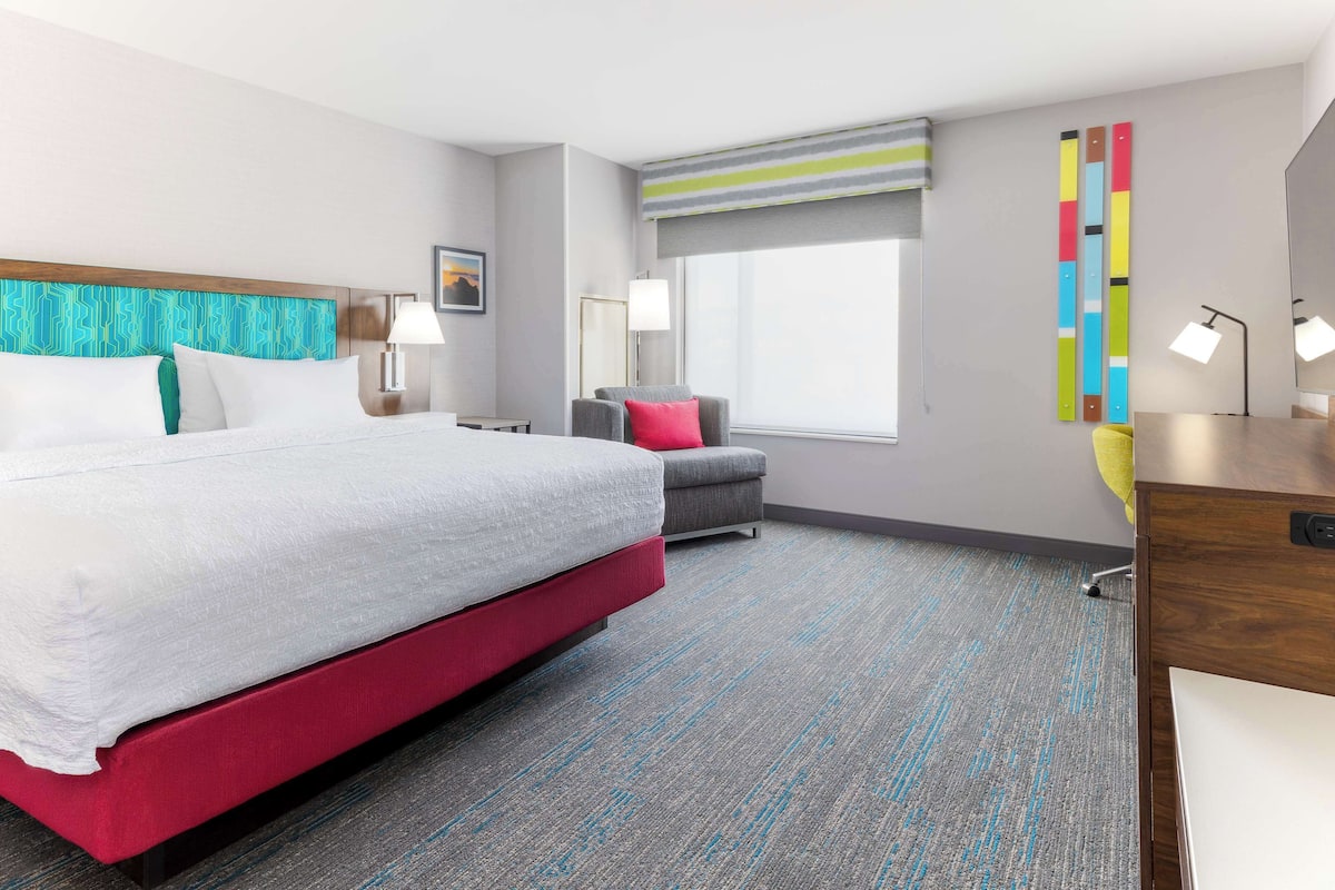 A modern hotel room with a large bed, a desk, a chair, and a window with a view.